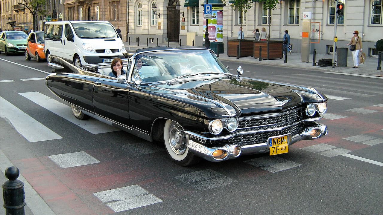 Black 1959 Cadillac Eldorado Biarritz convertible with chrome details and passengers driving through European city street intersection