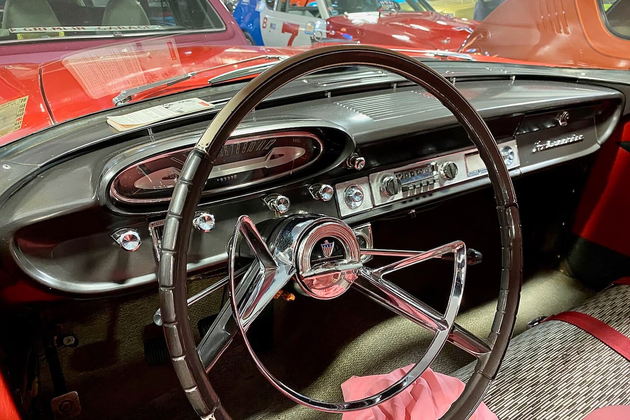 Vintage car interior showing chrome steering wheel, column-mounted manual transmission shifter, dashboard gauges, and red/green upholstery details