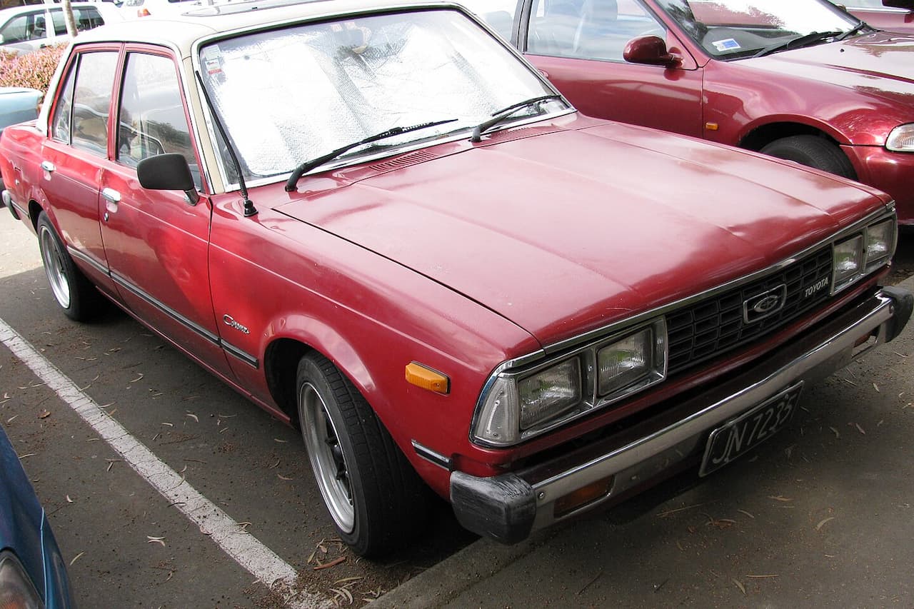 Red vintage Toyota sedan with reflective windshield sun shade parked in lot, other cars visible in background