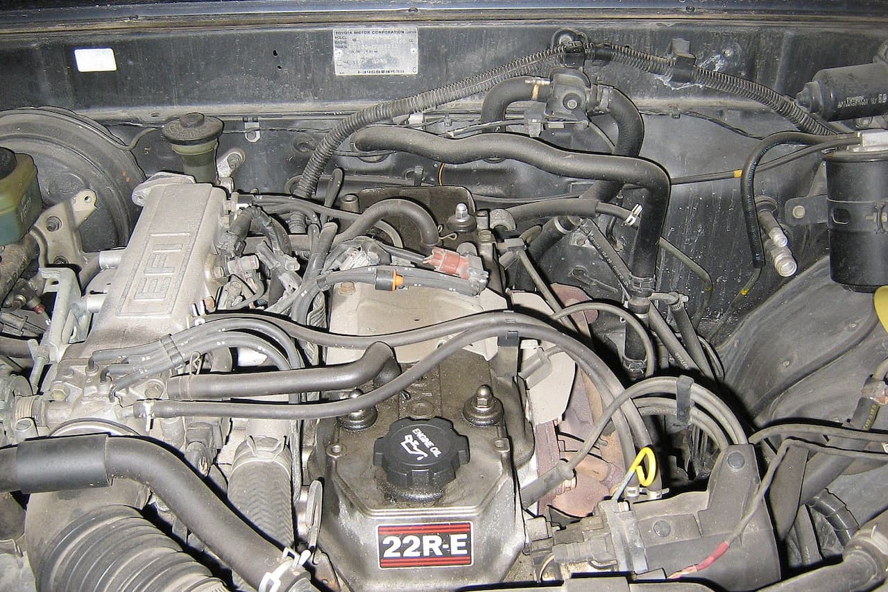 Toyota 22R-E engine bay showing carburetor, air cleaner, vacuum hoses, oil filler cap, and various engine components in weathered condition