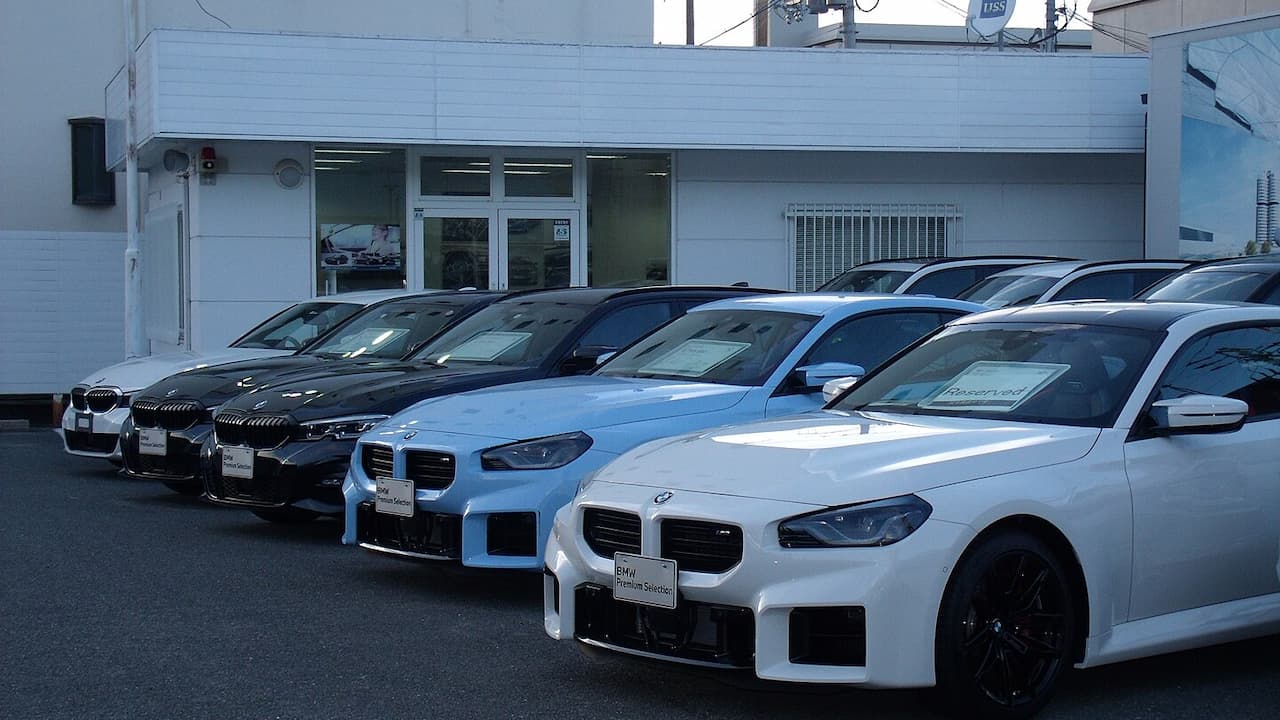 Row of used BMW cars parked at dealership lot with white building and glass showroom behind
