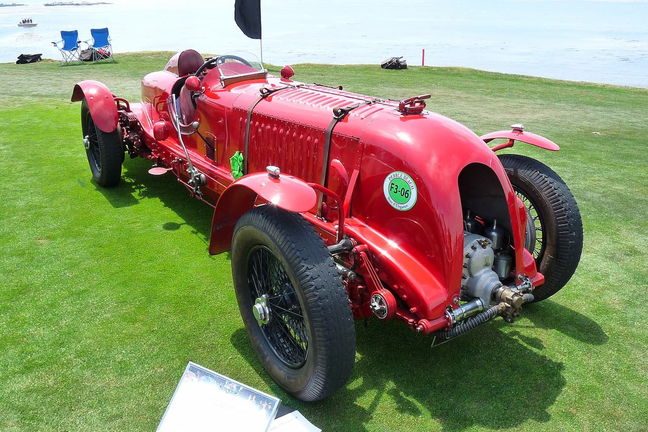 Red vintage Bentley Blower racing car displayed on green grass near waterfront with camping chairs and boat visible in background