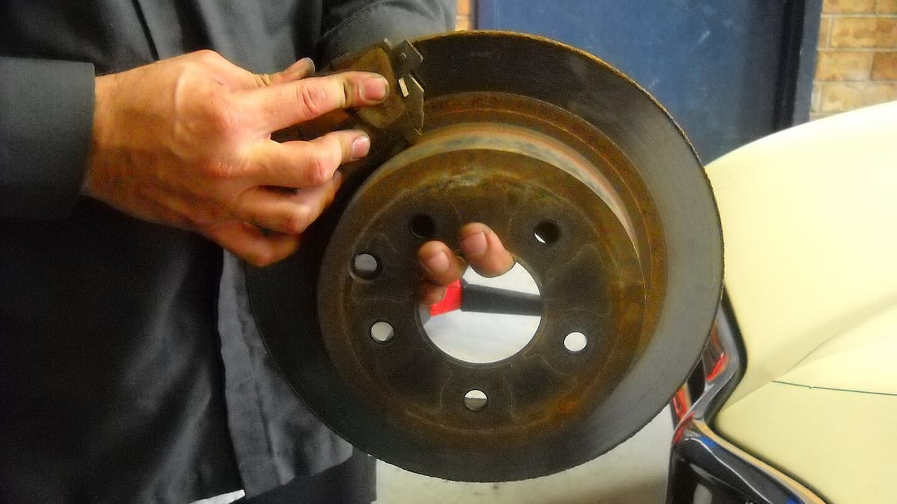 Person holding worn brake disc rotor showing brake pad material and bolt holes in automotive workshop setting