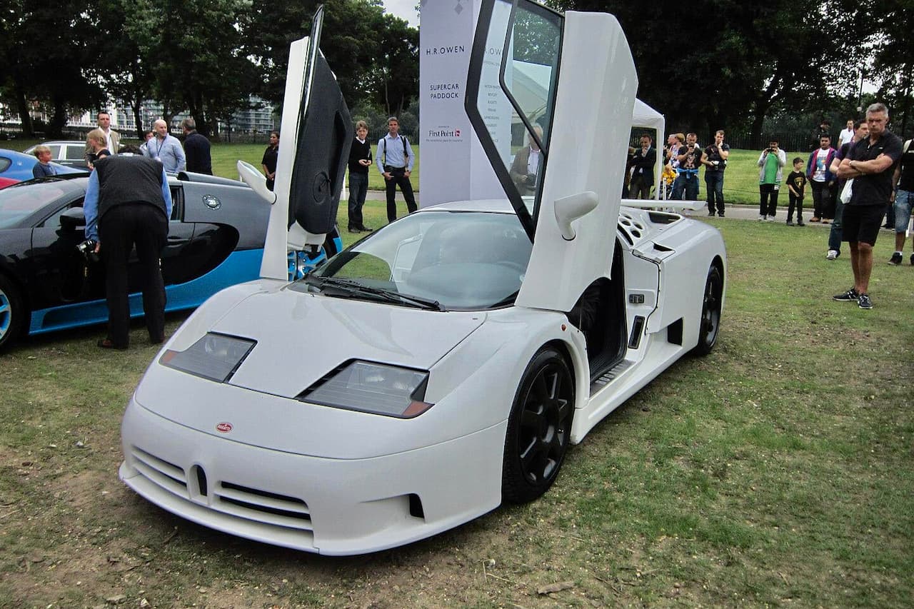 White Bugatti EB110 supercar with scissor doors open displayed at outdoor car show with spectators watching