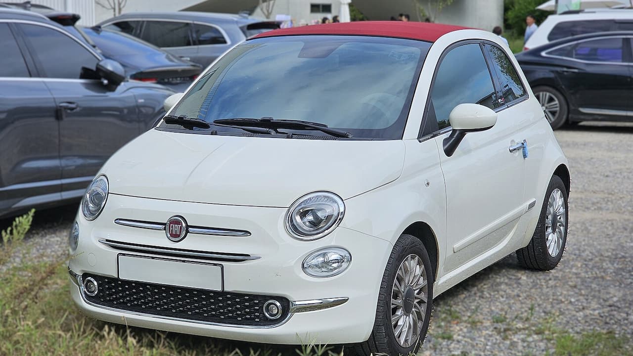 White Fiat 500C convertible with red soft top parked in gravel lot surrounded by other vehicles