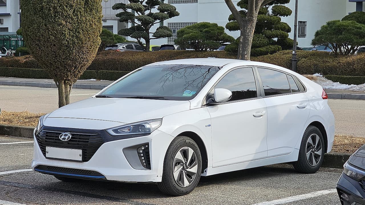 White 2025 Hyundai IONIQ Hybrid parked in urban lot with manicured trees and white buildings in background