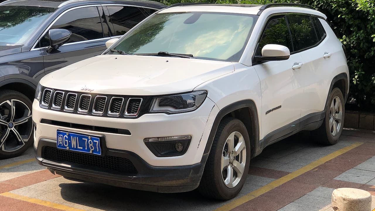 White Jeep Compass SUV with Chinese license plate parked in lot next to dark sedan and greenery
