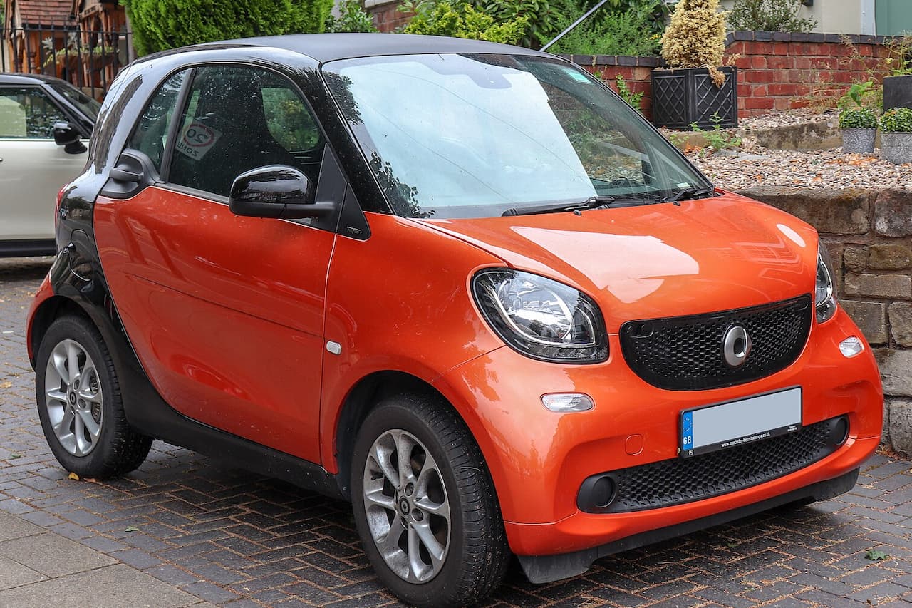Orange and black Smart ForTwo car parked on cobblestone street with brick wall and greenery in background