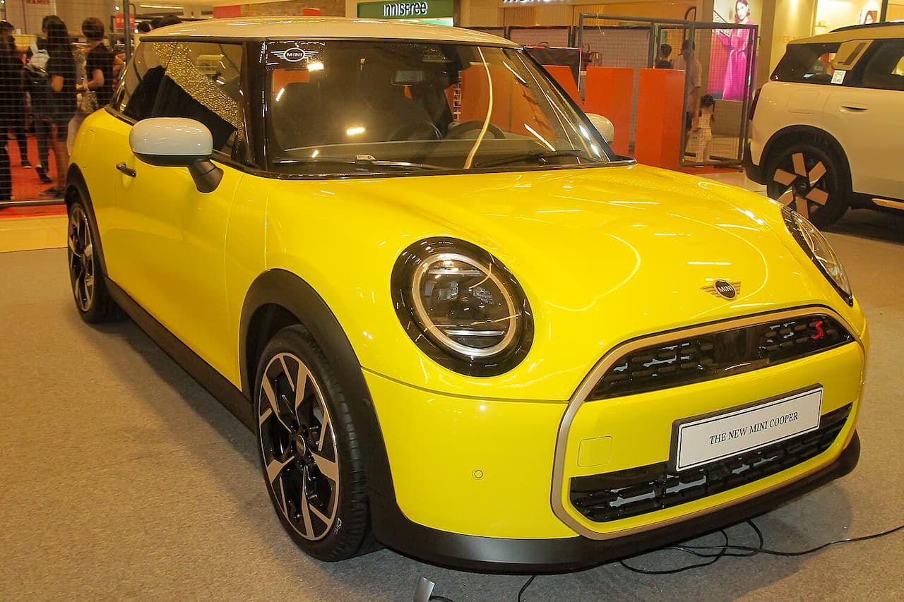 Bright yellow 2025 Mini Cooper Electric displayed at auto show with black roof and distinctive round headlights