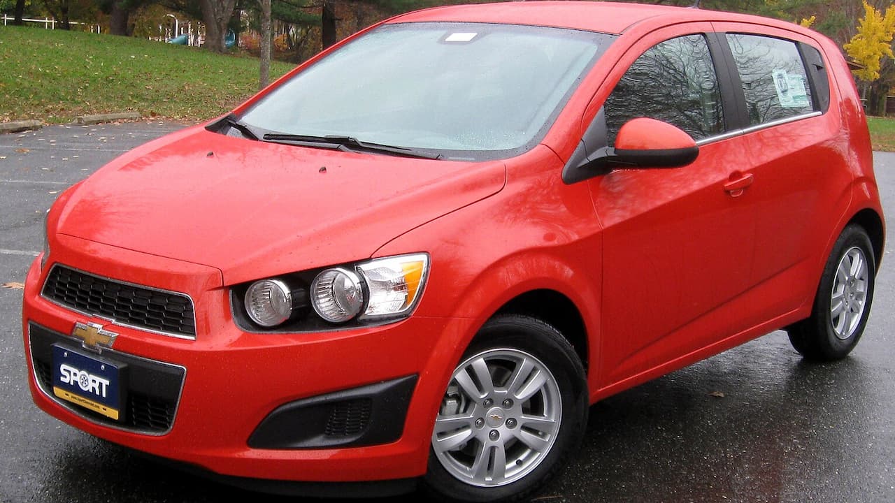 Red Chevrolet Sonic parked on wet asphalt with dual round headlights and alloy wheels visible