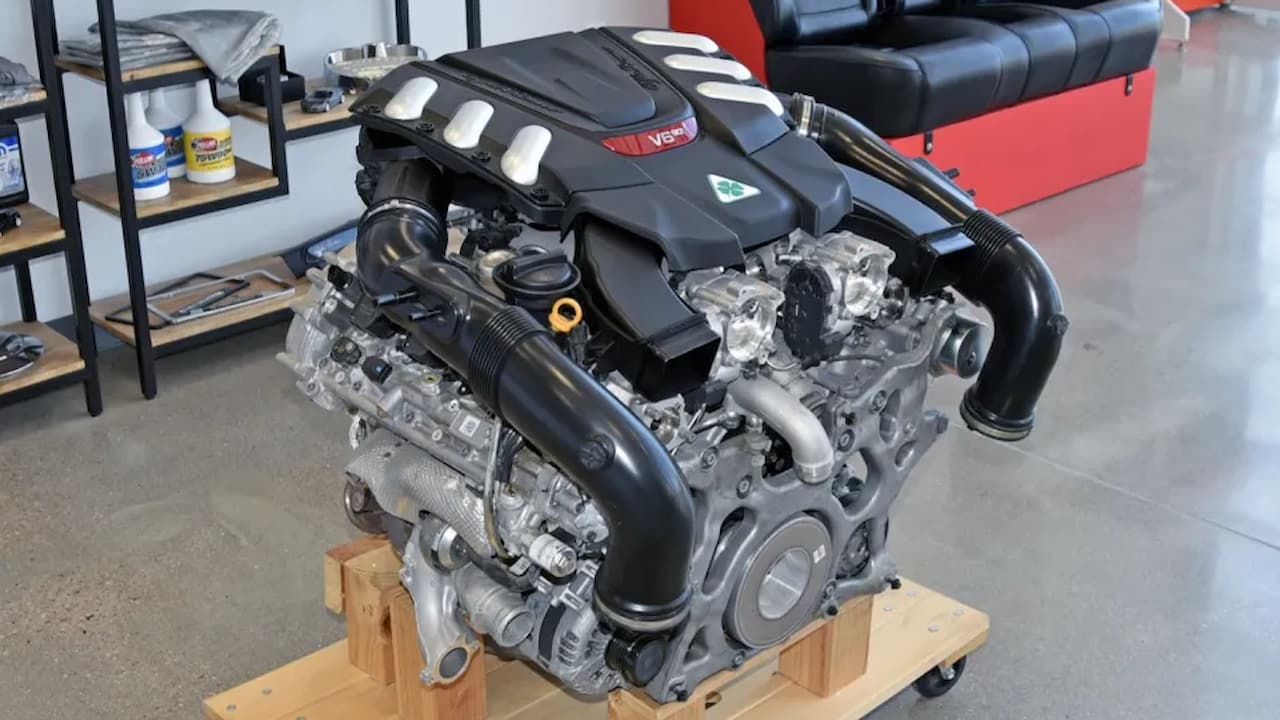 Alfa Romeo 2.9L V6 Bi-Turbo engine on wooden stand showing black plastic cover, dual turbochargers, and complex intake system