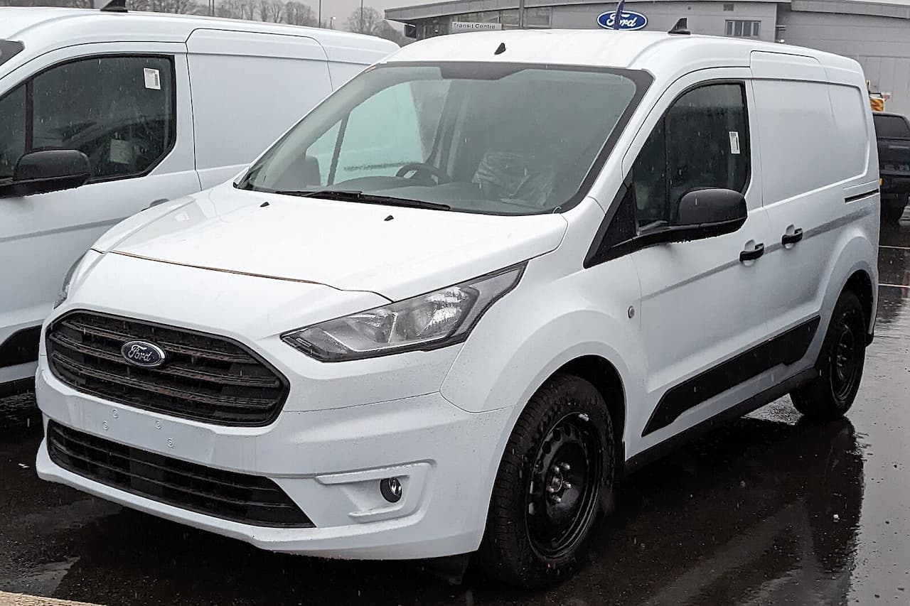White 2022 Ford Transit Connect cargo van with black wheels parked at Ford dealership on wet pavement