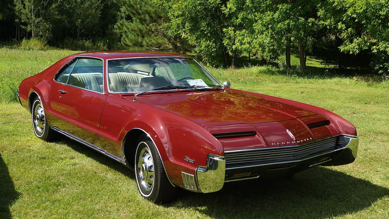 Red 1966 Oldsmobile Toronado classic car with distinctive front grille parked on grass beside wooded area