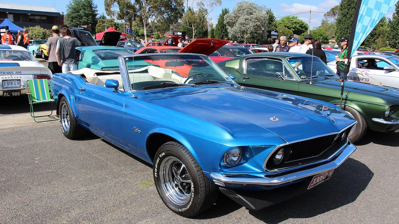 Blue 1969 Ford Mustang convertible with white soft top and chrome wheels at outdoor classic car show