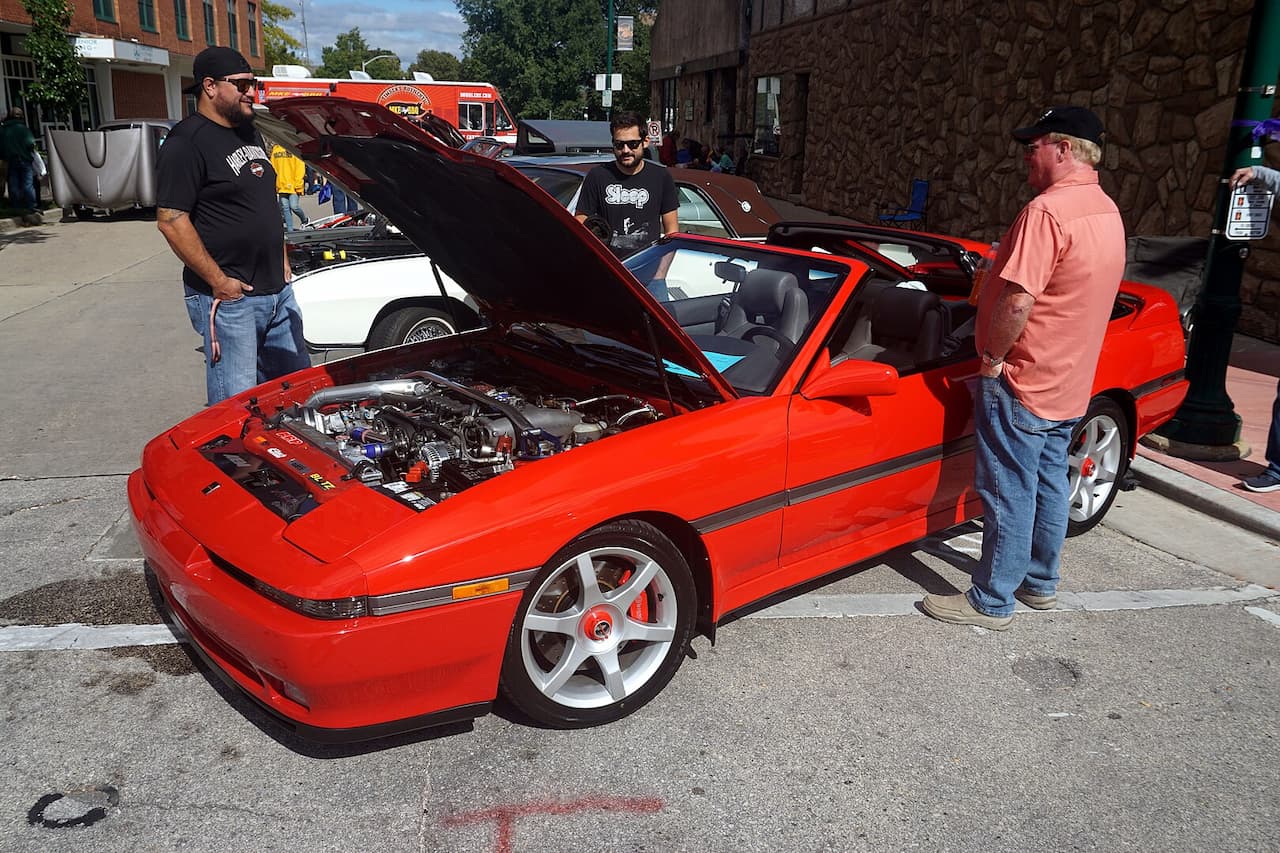Red sports car with hood open displaying engine at street car show, with three men examining the vehicle