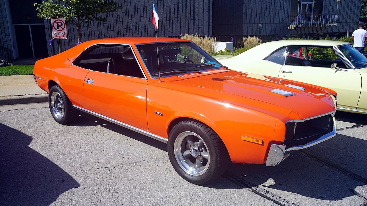 Orange 1970 AMC Javelin muscle car parked at car show with chrome wheels, next to cream-colored classic car