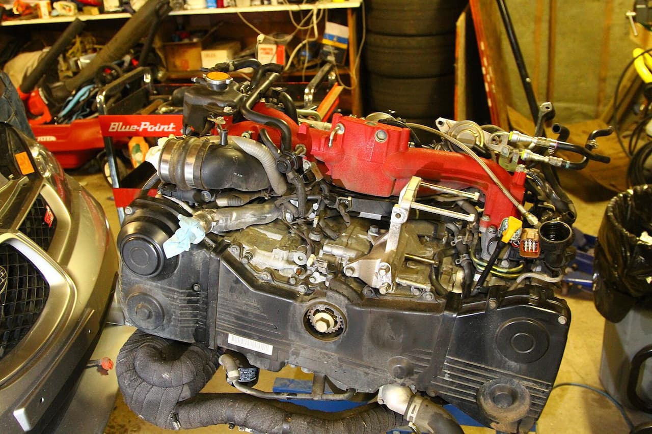 Disassembled Subaru EJ25 engine on workbench showing red cylinder heads, black crankcase, and various mechanical components during rebuild