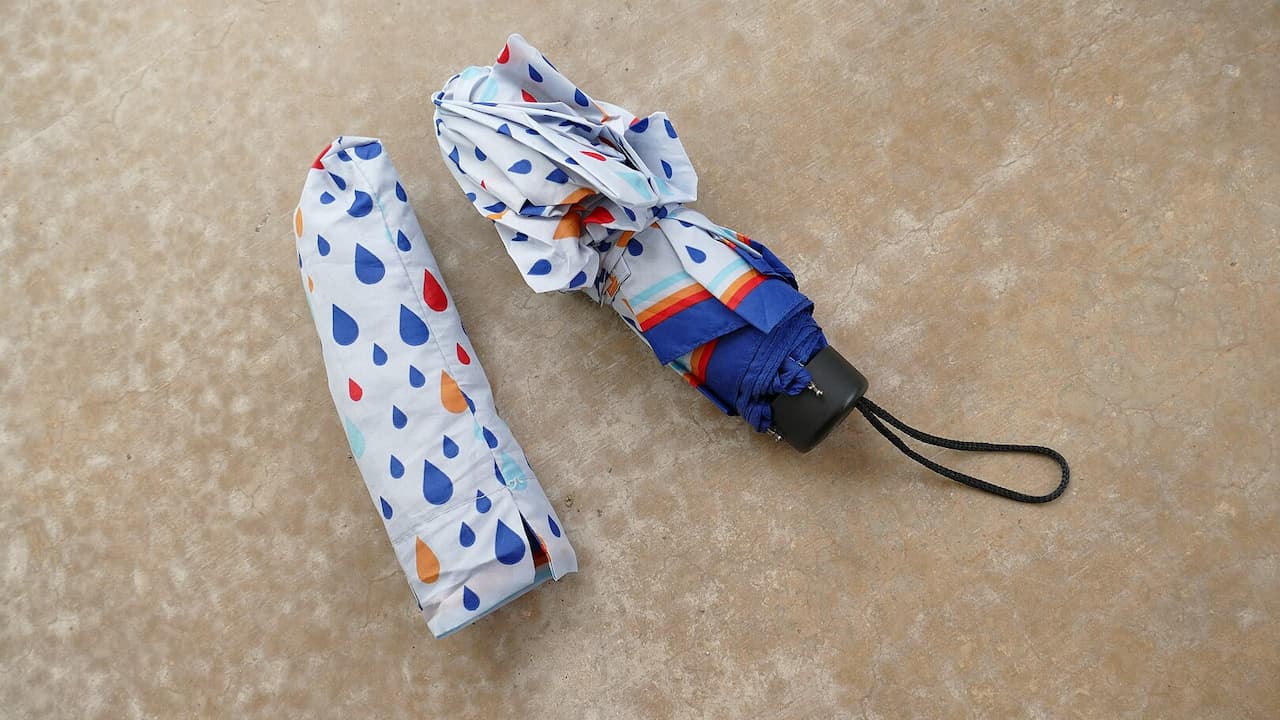 A compact umbrella with colorful raindrop pattern lies folded next to its matching storage sleeve on beige surface