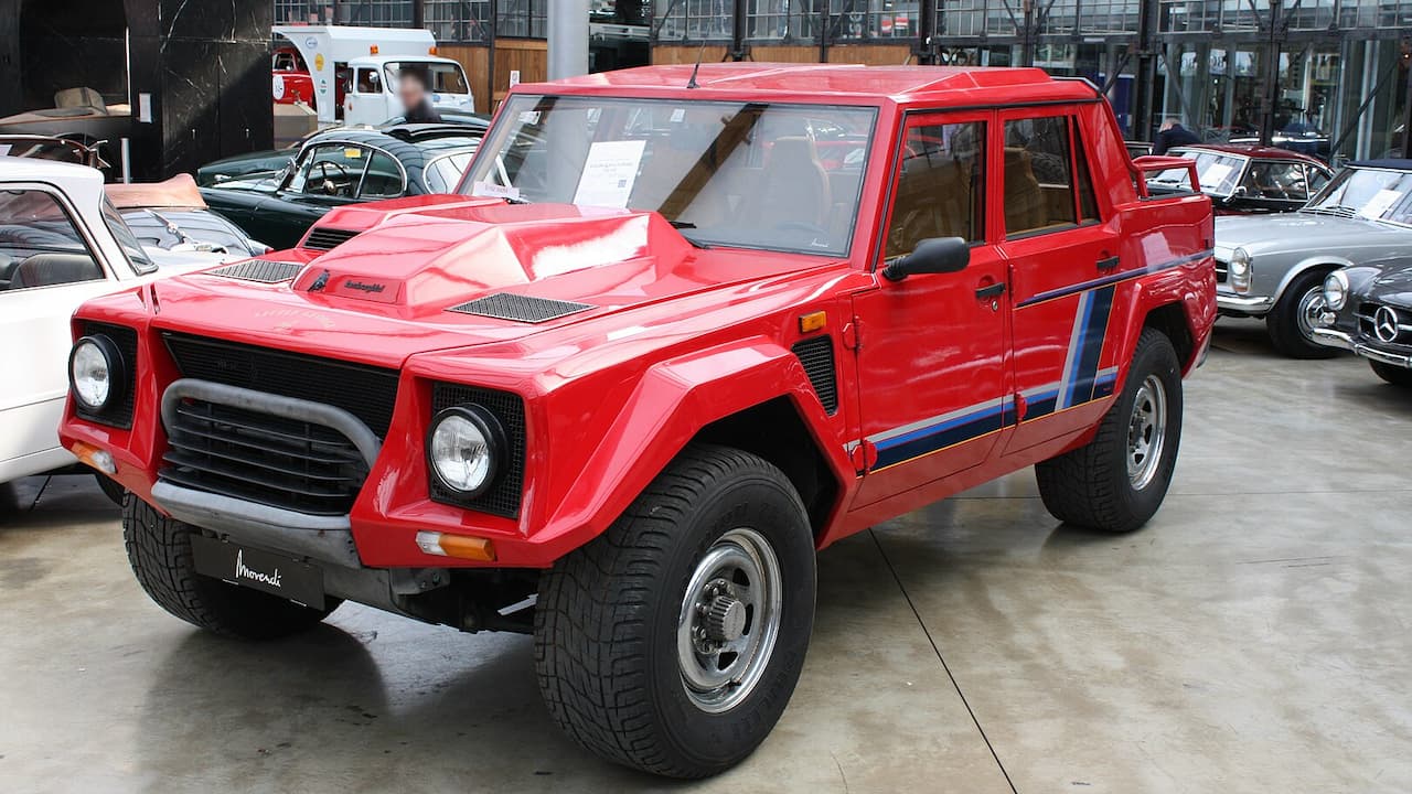 Red Lamborghini LM002 SUV with blue racing stripes displayed in showroom alongside classic cars and vintage vehicles