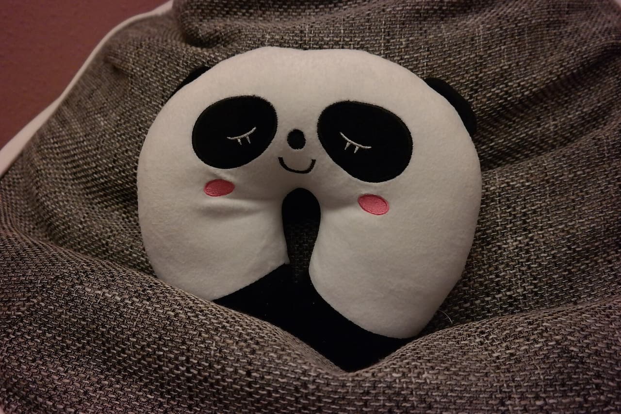 White panda-shaped travel neck pillow with black eye patches, pink cheeks, and sleepy expression on gray fabric