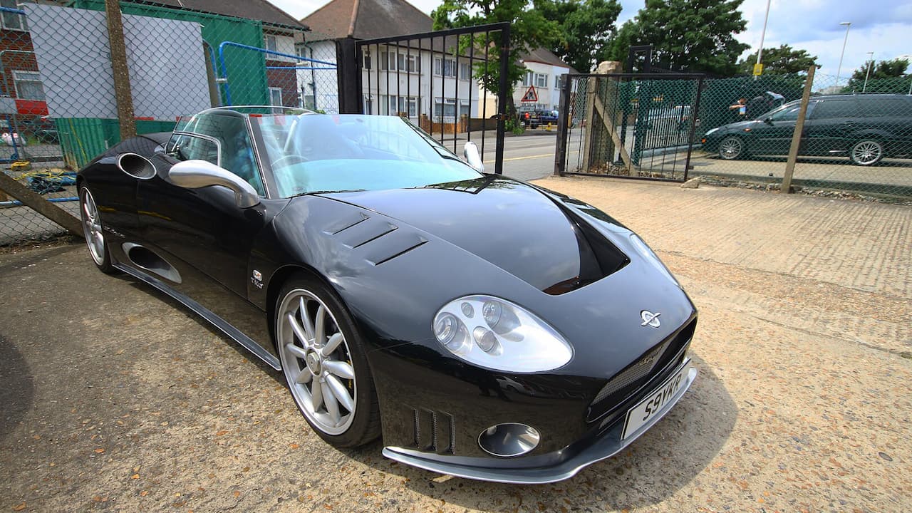 Black Spyker C8 sports car with distinctive headlights and alloy wheels parked on gravel surface