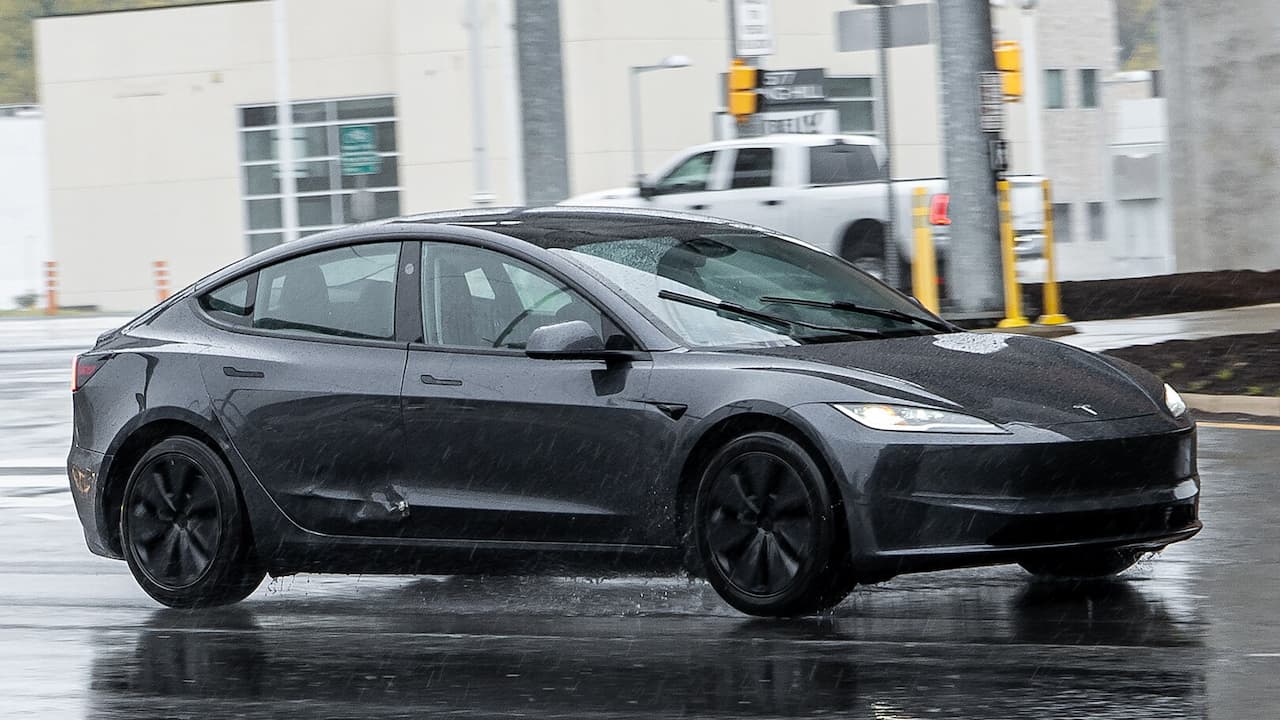 Dark gray 2025 Tesla Model 3 driving on wet urban street with buildings and traffic lights visible