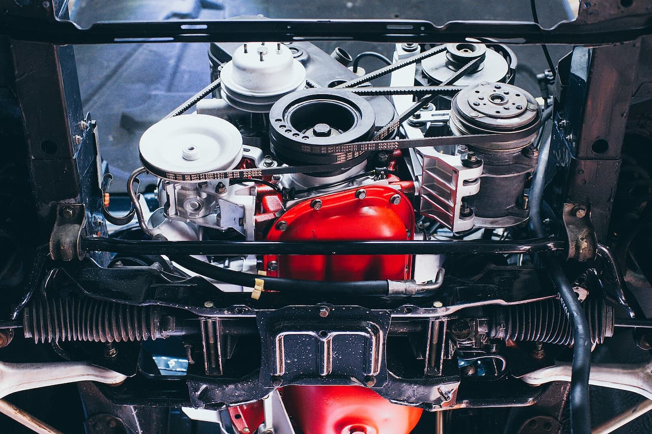 Volvo B230F engine bay viewed from below showing red engine block, pulleys, belts, air cleaner, and various mechanical components underneath