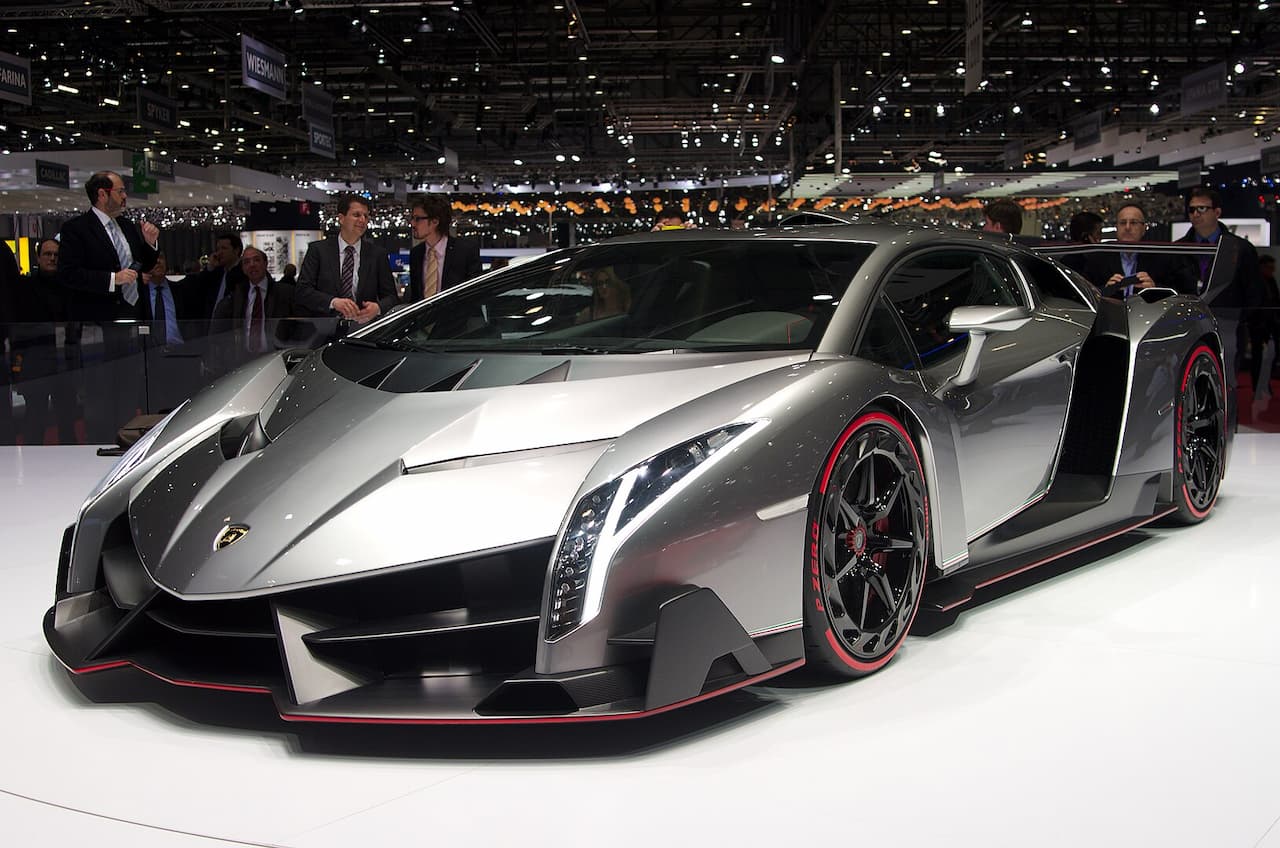 Gray Lamborghini Veneno supercar with red accents displayed at auto show with crowds and exhibition lighting in background