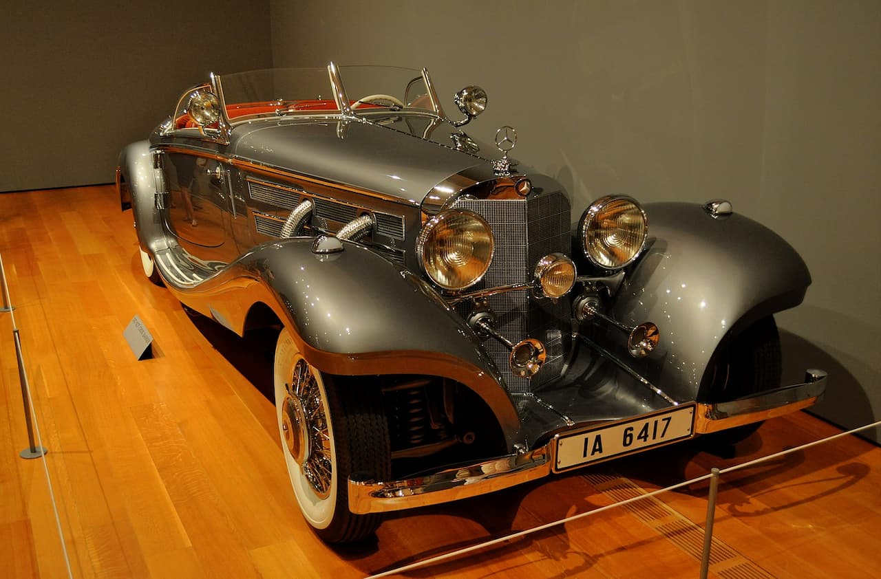 Gray Mercedes-Benz 540K Special Roadster classic car with multiple headlights and wire wheels displayed in museum on wooden platform