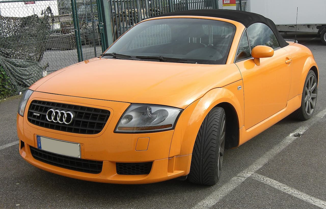 Bright orange Audi TT Roadster convertible with black soft top parked on street near fencing