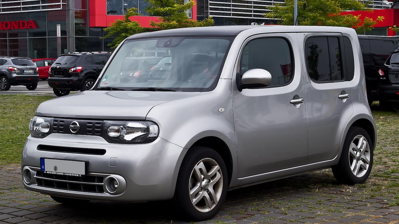 Silver Nissan Cube compact van with distinctive boxy design and round headlights parked on cobblestone near Honda dealership