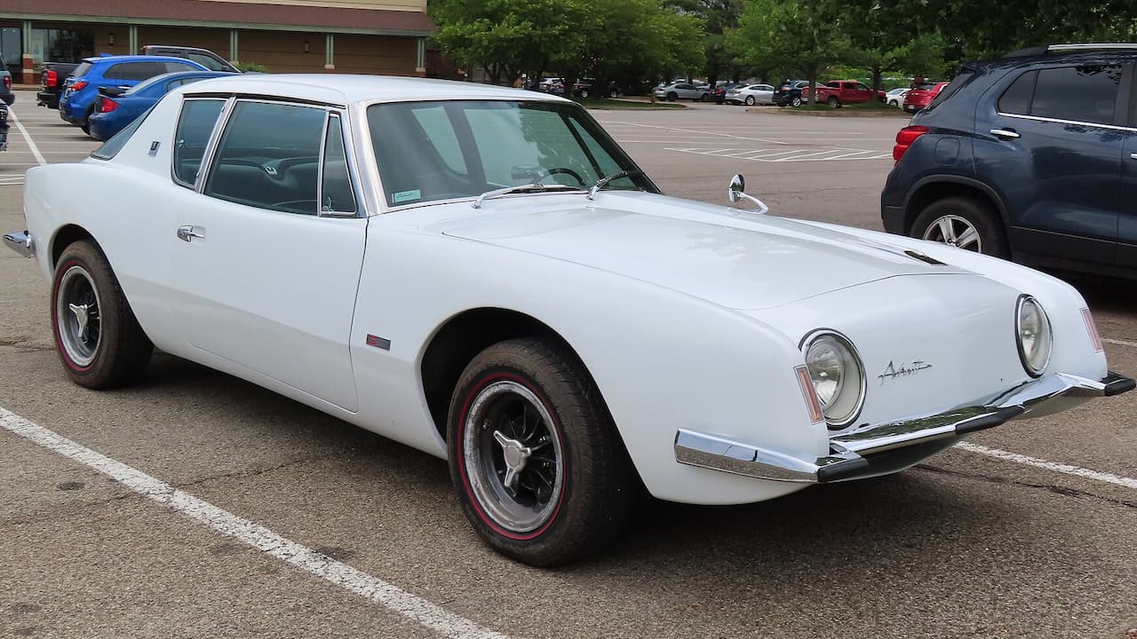 White 1963 Studebaker Avanti sports car with distinctive round headlights and red-striped tires in parking lot