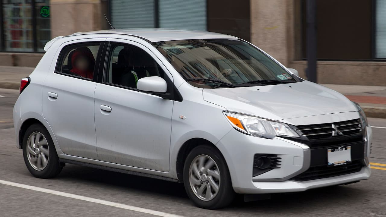 White Mitsubishi Mirage driving on city street with passengers visible through windows, urban buildings in background