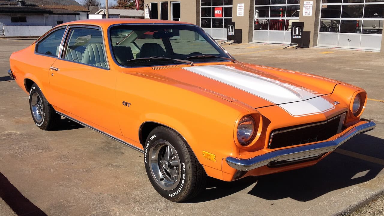 Orange Chevrolet Vega classic muscle car with white racing stripes parked outside commercial building storefront