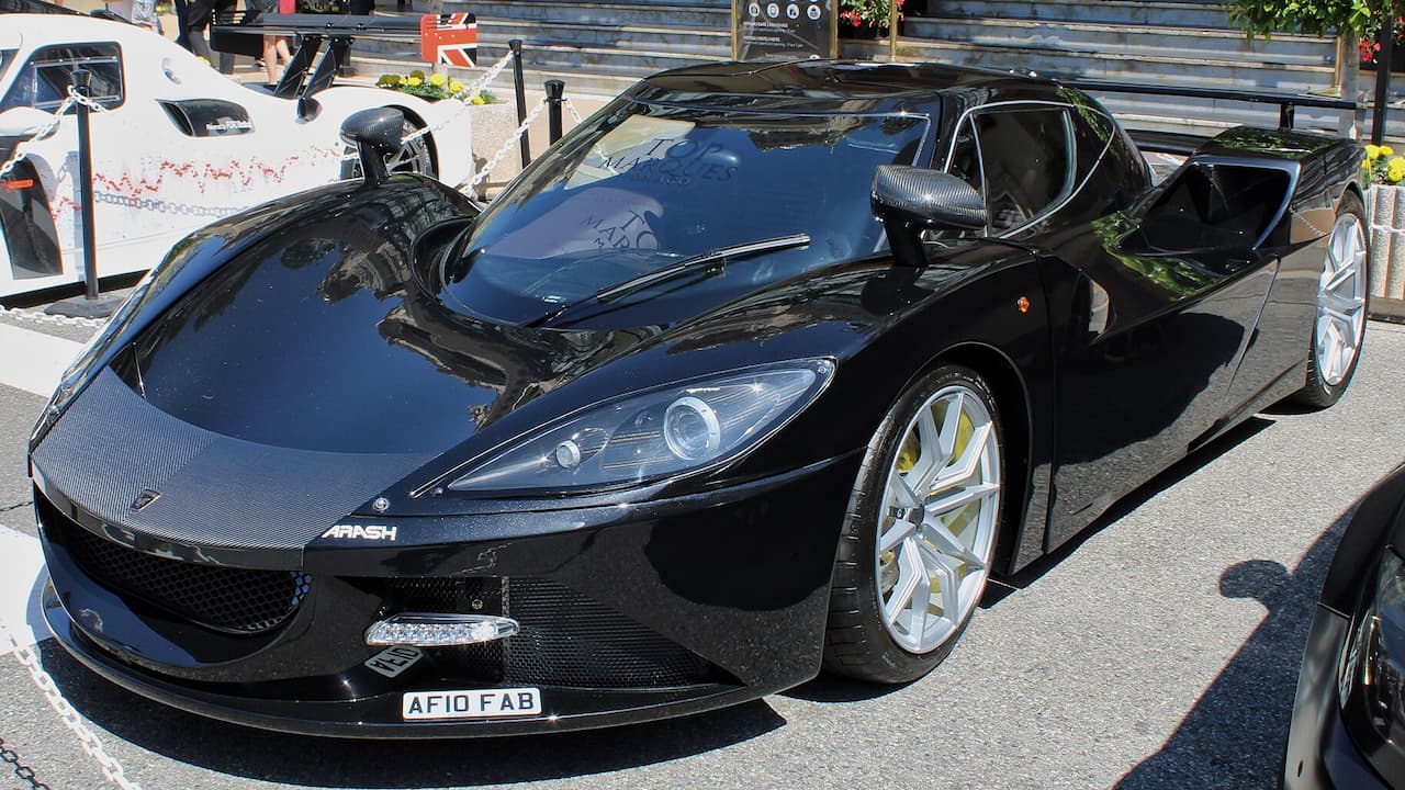 Black Arash AF10 supercar with carbon fiber bodywork and white wheels parked at outdoor car show