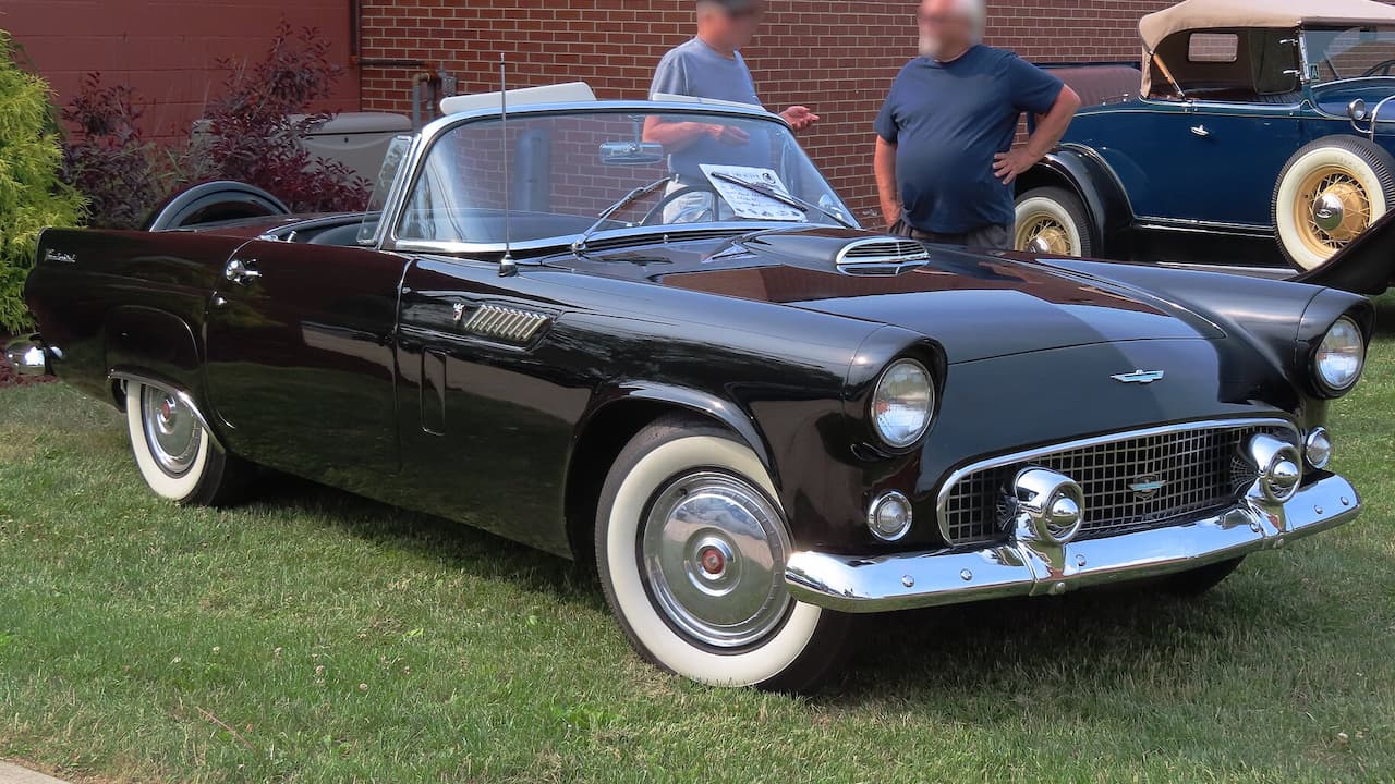 Black 1956 Ford Thunderbird convertible with white hardtop and whitewall tires parked on grass at car show