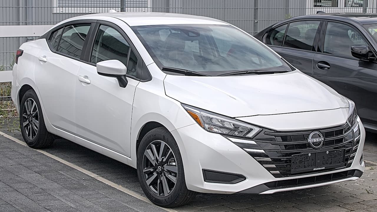 White Nissan Versa sedan parked in lot with distinctive V-motion grille, LED headlights, and dark alloy wheels