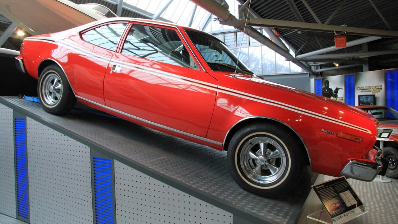 Red AMC Hornet X hatchback with white racing stripes displayed on platform in modern automotive museum with glass ceiling