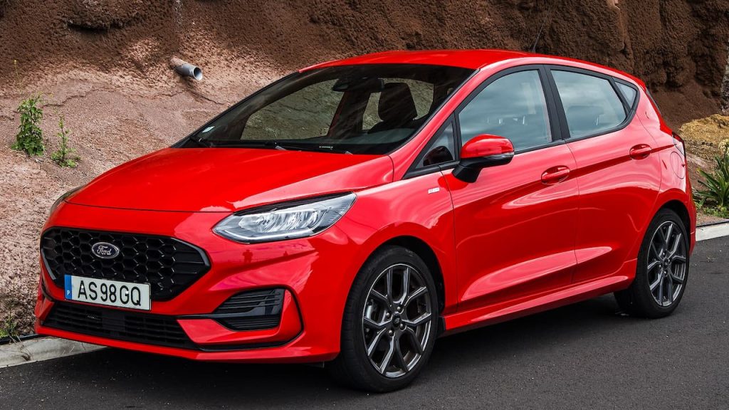 Red Ford Fiesta with black grille and alloy wheels parked on asphalt road beside dirt embankment