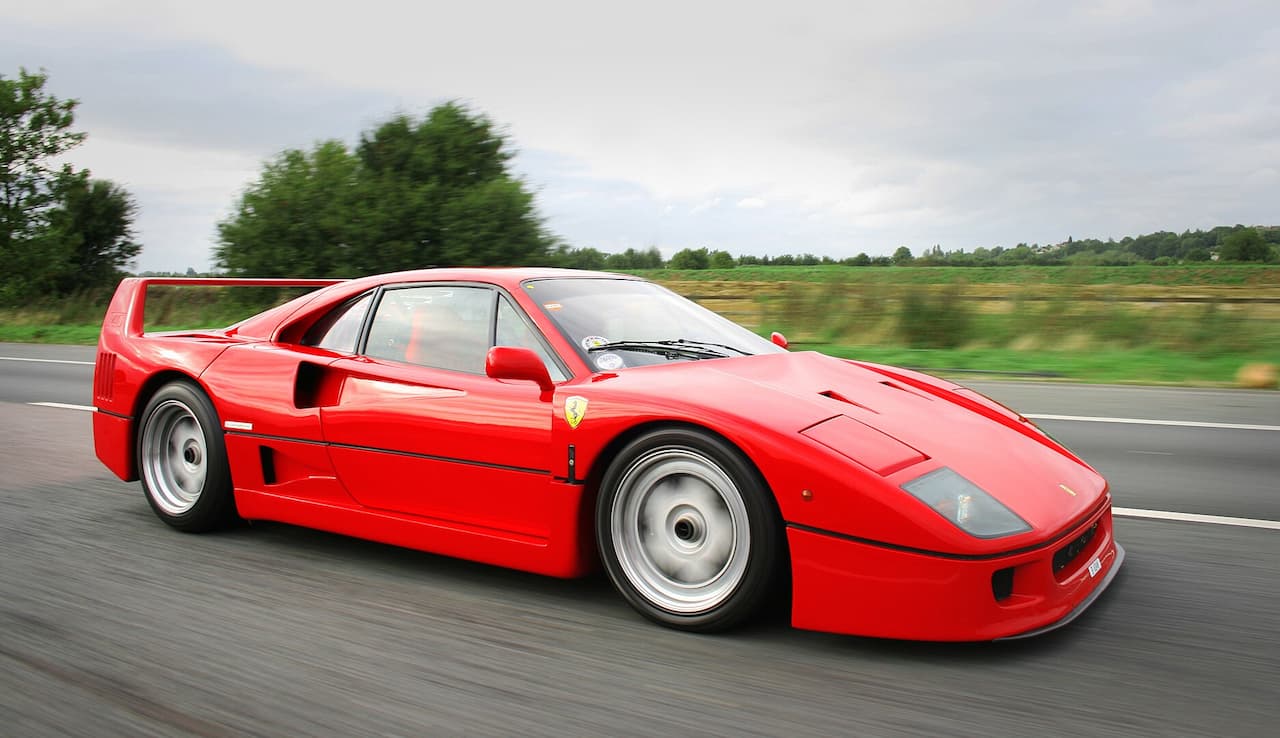 Red Ferrari F40 supercar with distinctive rear wing and side air intakes driving on highway with countryside background
