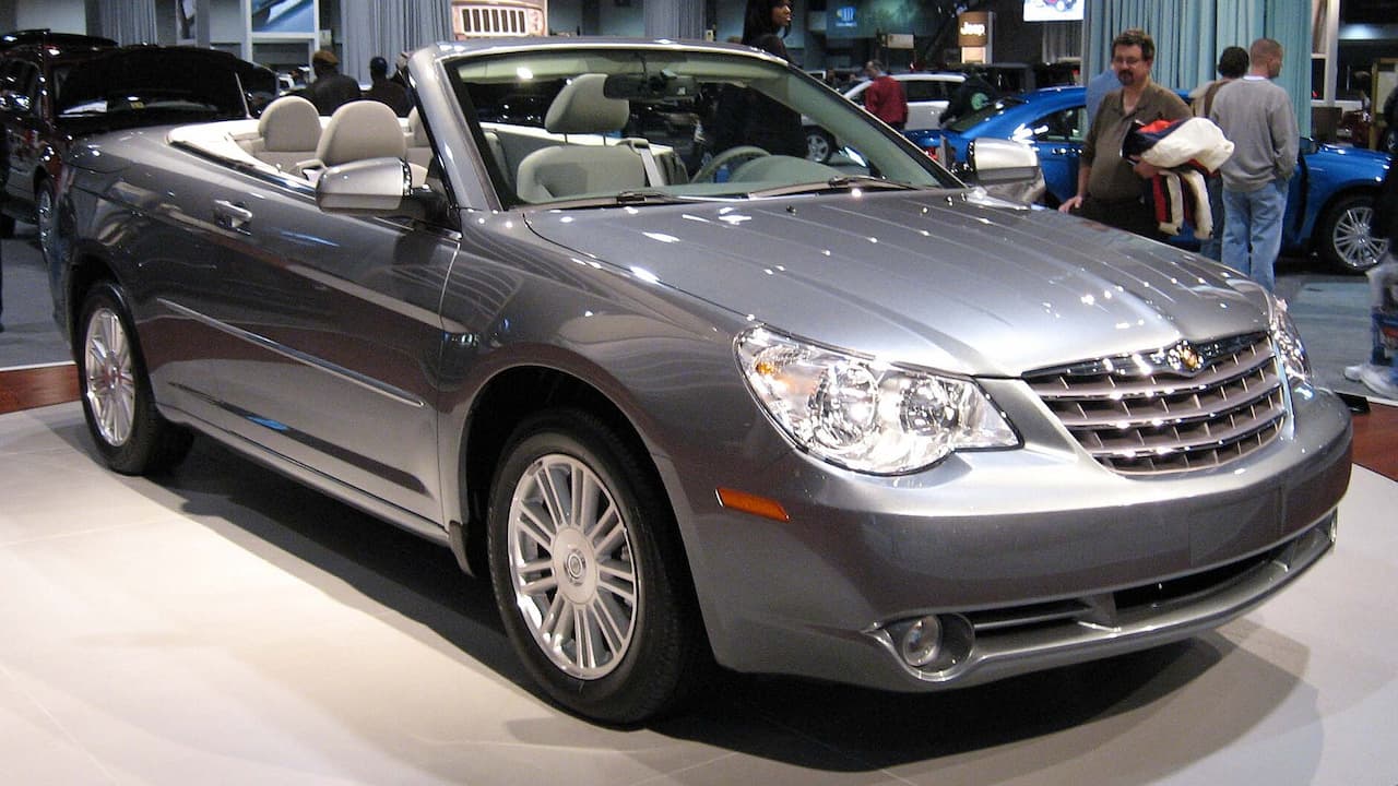 Silver Chrysler Sebring convertible with beige leather interior and multi-spoke wheels displayed at indoor auto show with visitors