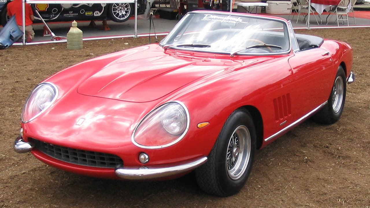 Red 1967 Ferrari 275 GTB/4 NART Spider convertible with wire wheels displayed at racing event with pit area background