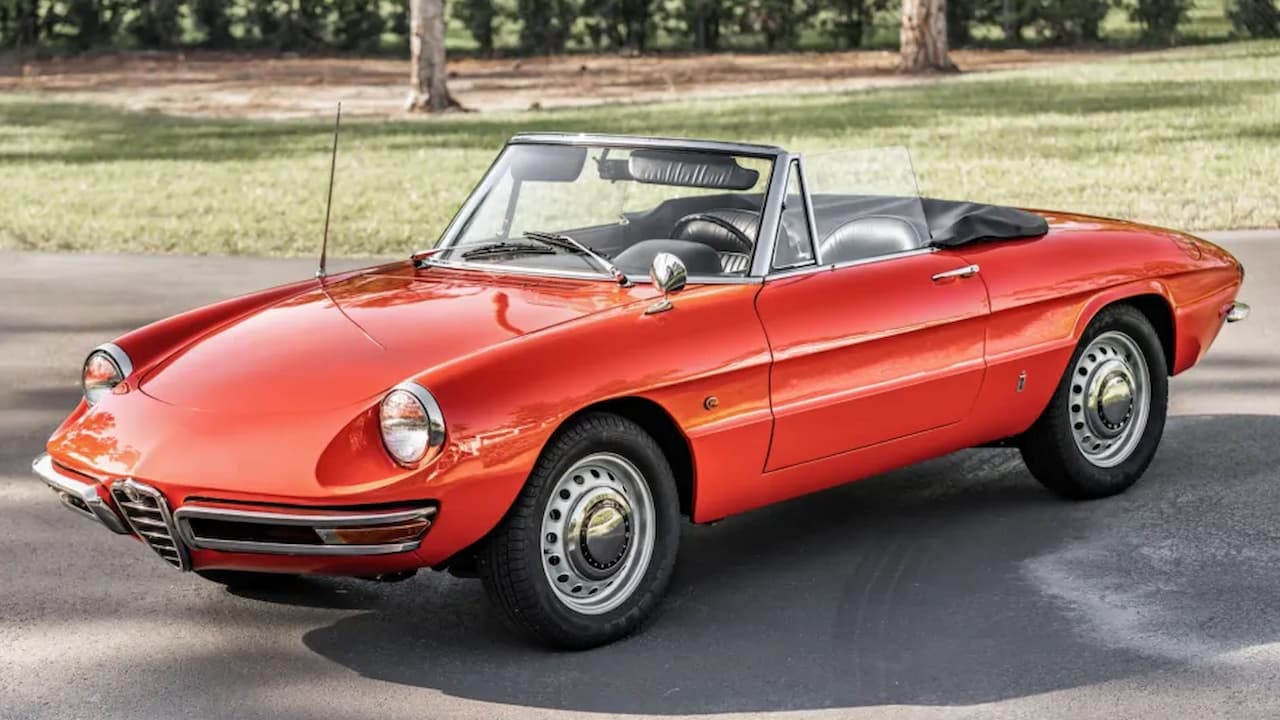 Red 1966 Alfa Romeo Spider Duetto parked on asphalt road with trees and grass in background