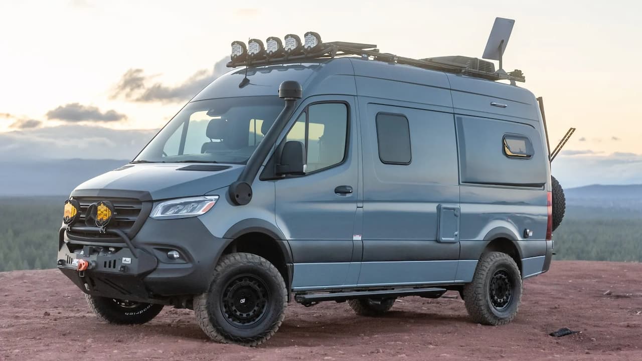 Gray Winnebago Revel adventure van with roof rack, off-road tires, and accessories parked on desert terrain