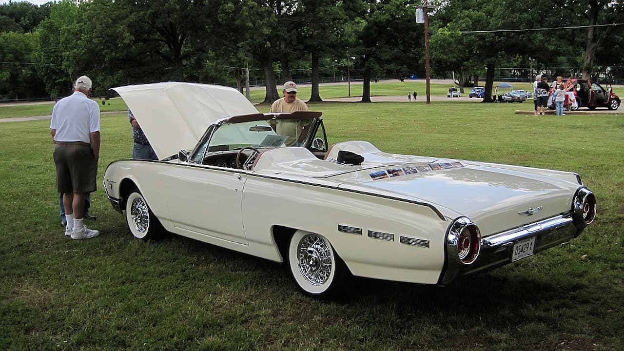 White classic convertible car with soft top at outdoor car show, two men examining vehicle on grass field
