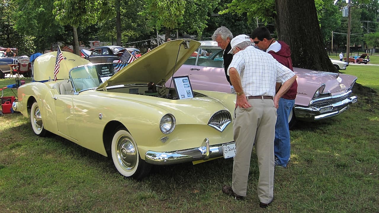 Yellow vintage convertible with hood open at car show, three men examining engine, other classic cars visible in background