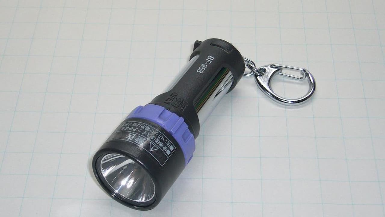 Black flashlight with blue accent ring and metal keychain attachment on white grid paper background