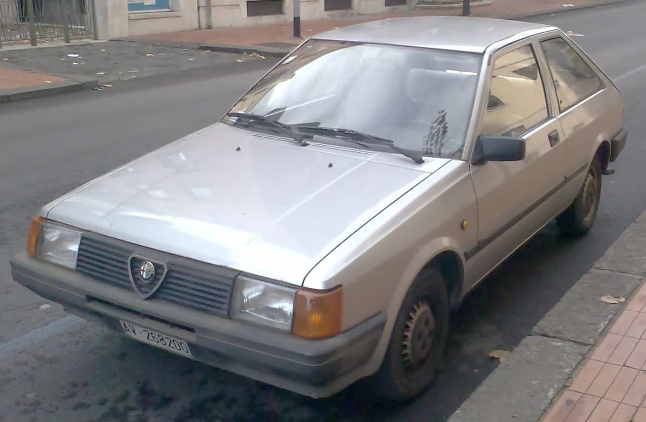 Beige Alfa Romeo Arna hatchback parked on street with distinctive grille, orange turn signals, and steel wheels visible