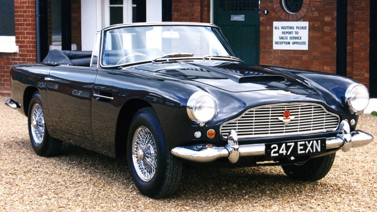 Dark blue 1961 Aston Martin DB4 GT Zagato convertible with wire wheels and UK license plate parked outside brick building