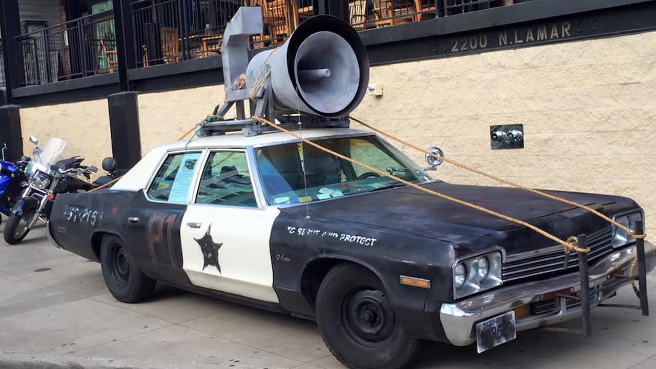 Weathered black and white police car with large megaphone on roof, styled as the Bluesmobile from Blues Brothers movie